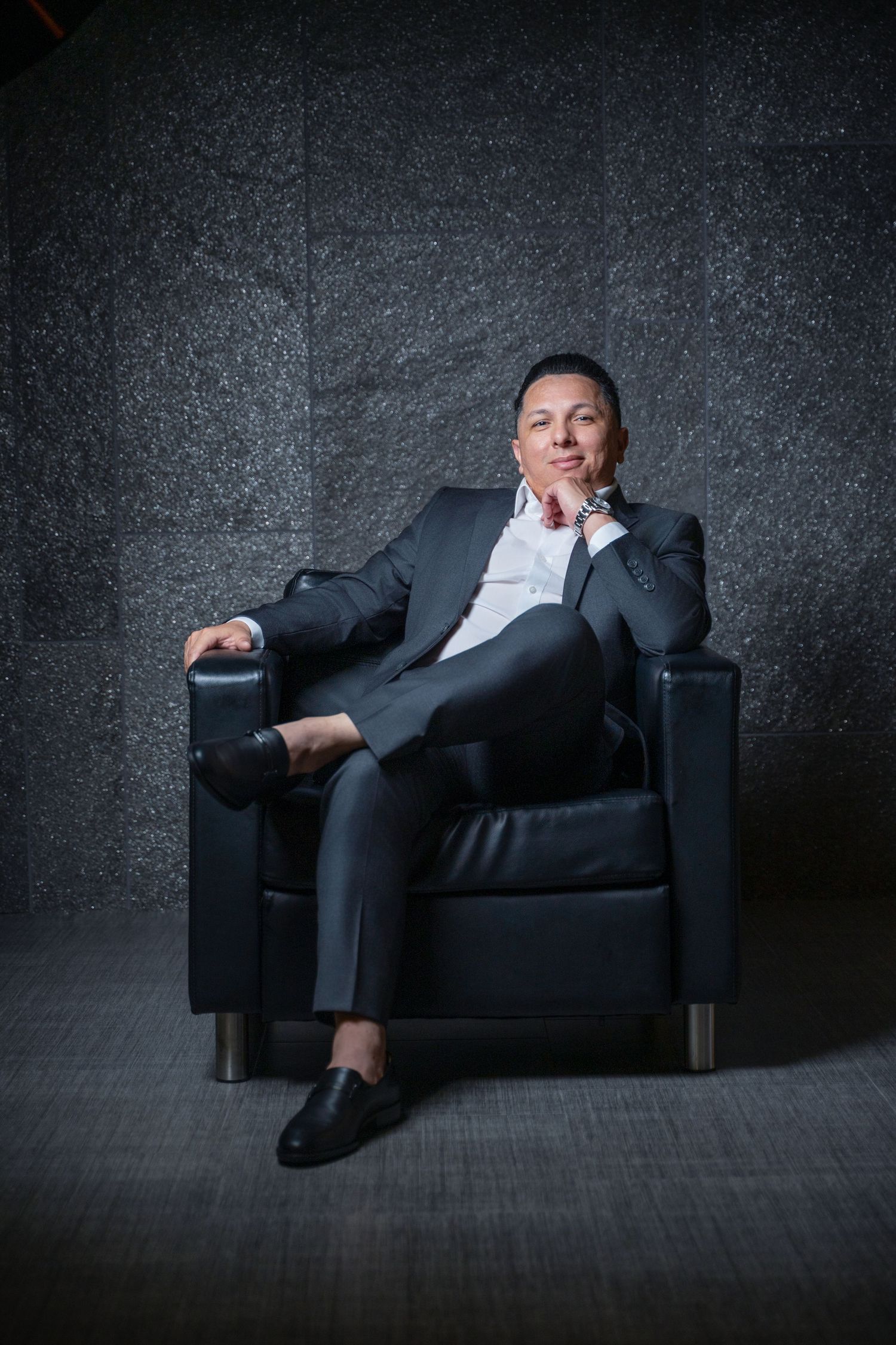 Professional portrait of someone seated in a black leather armchair against a dark textured background wearing a suit.