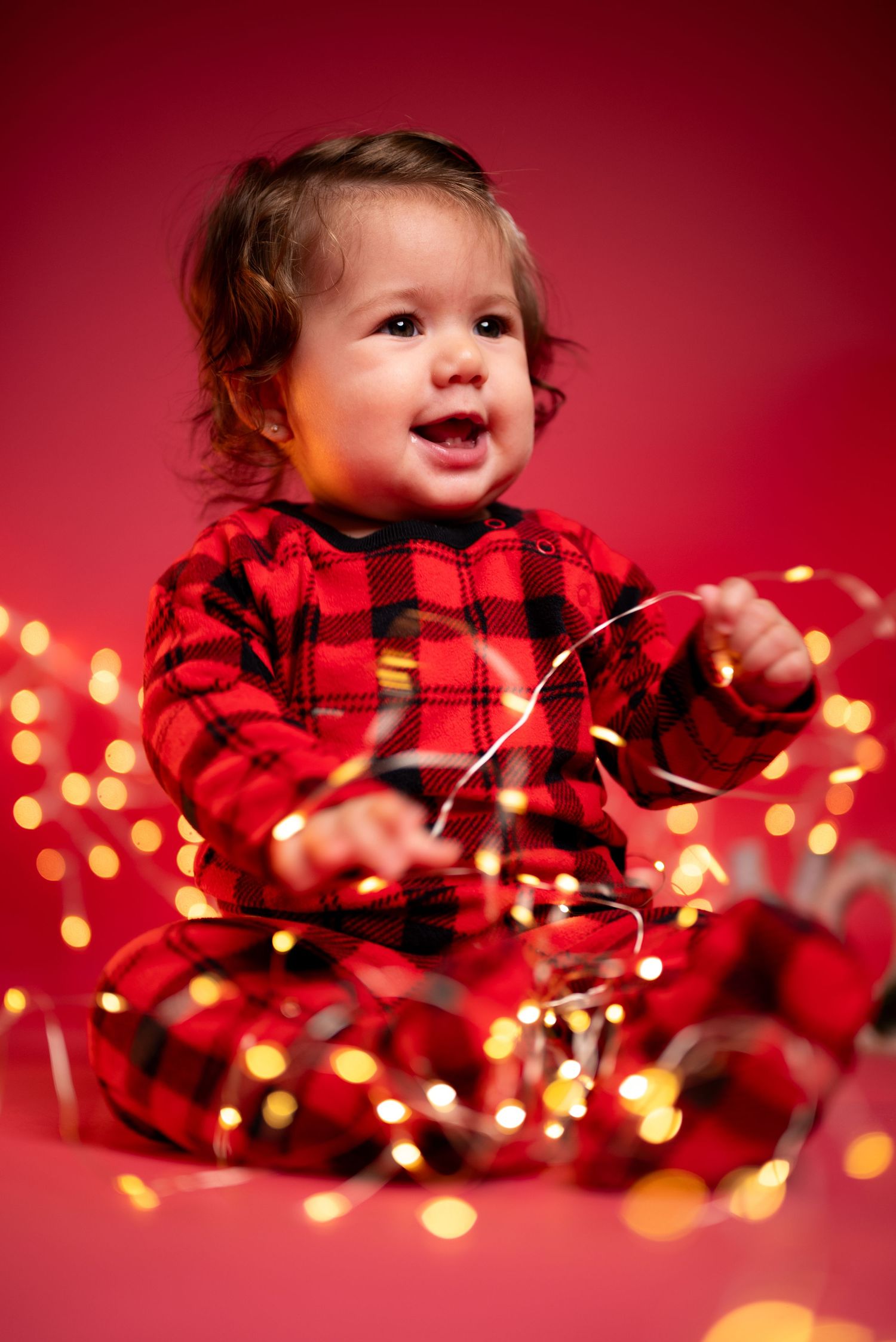 Christmas-themed baby portrait taken in Houston, Texas, featuring a baby in festive pajamas wrapped in LED string lights against a bright re