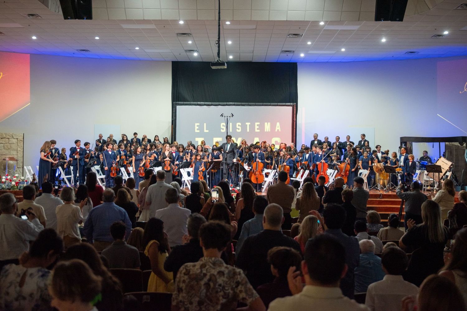 A large choir performs on stage in an auditorium with blue accent lighting while an audience stands watching.