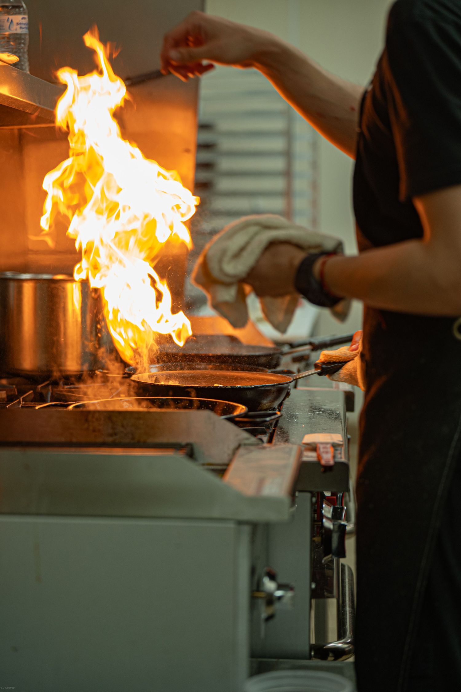 Professional kitchen cooking scene with dramatic flames rising from stovetop during food preparation.