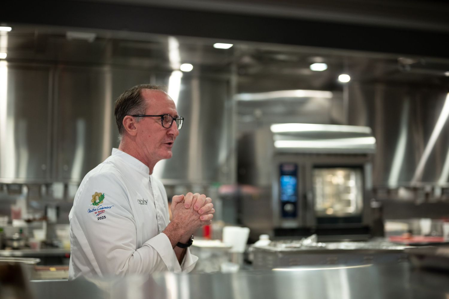 Professional chef in white uniform stands in modern commercial kitchen with stainless steel equipment.