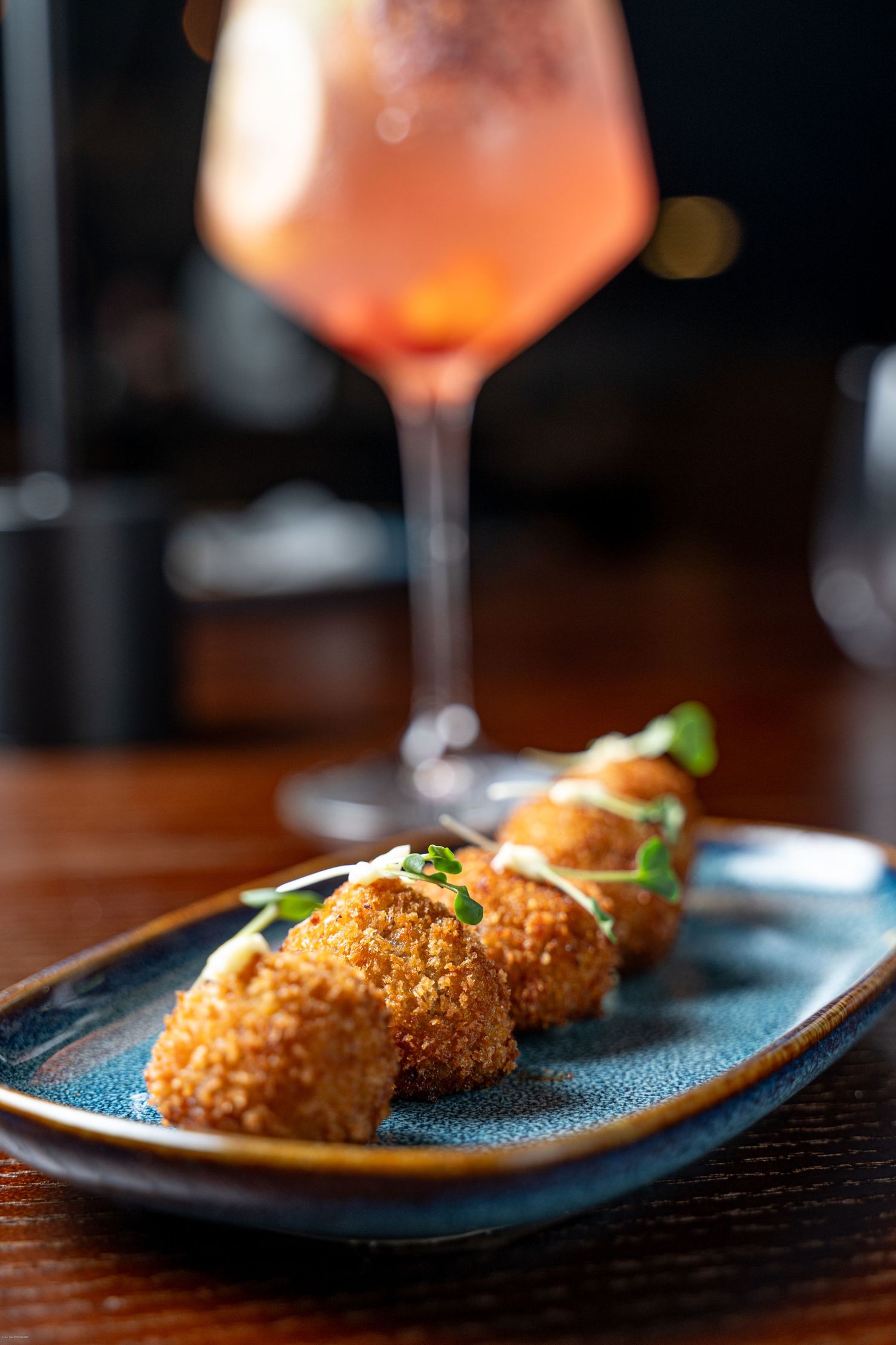 Crispy golden breaded croquettes served on a modern black plate with cocktails in the background.