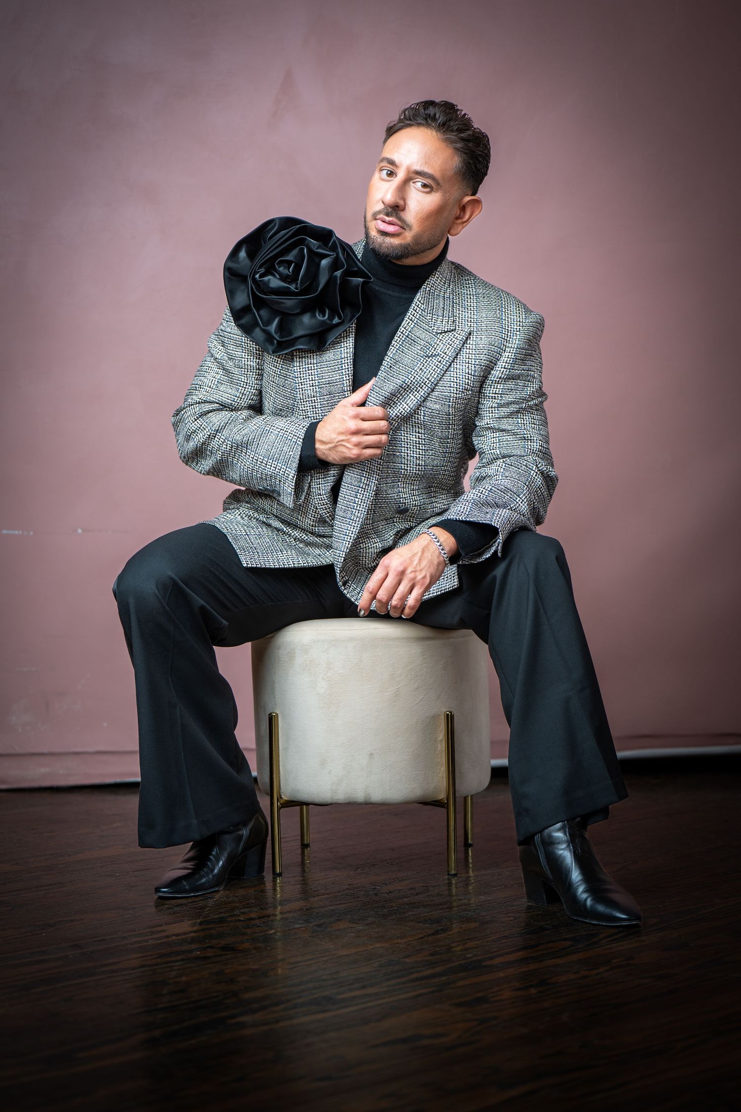 Person in gray blazer and black pants sits on white chair against pink wall in moody portrait series.