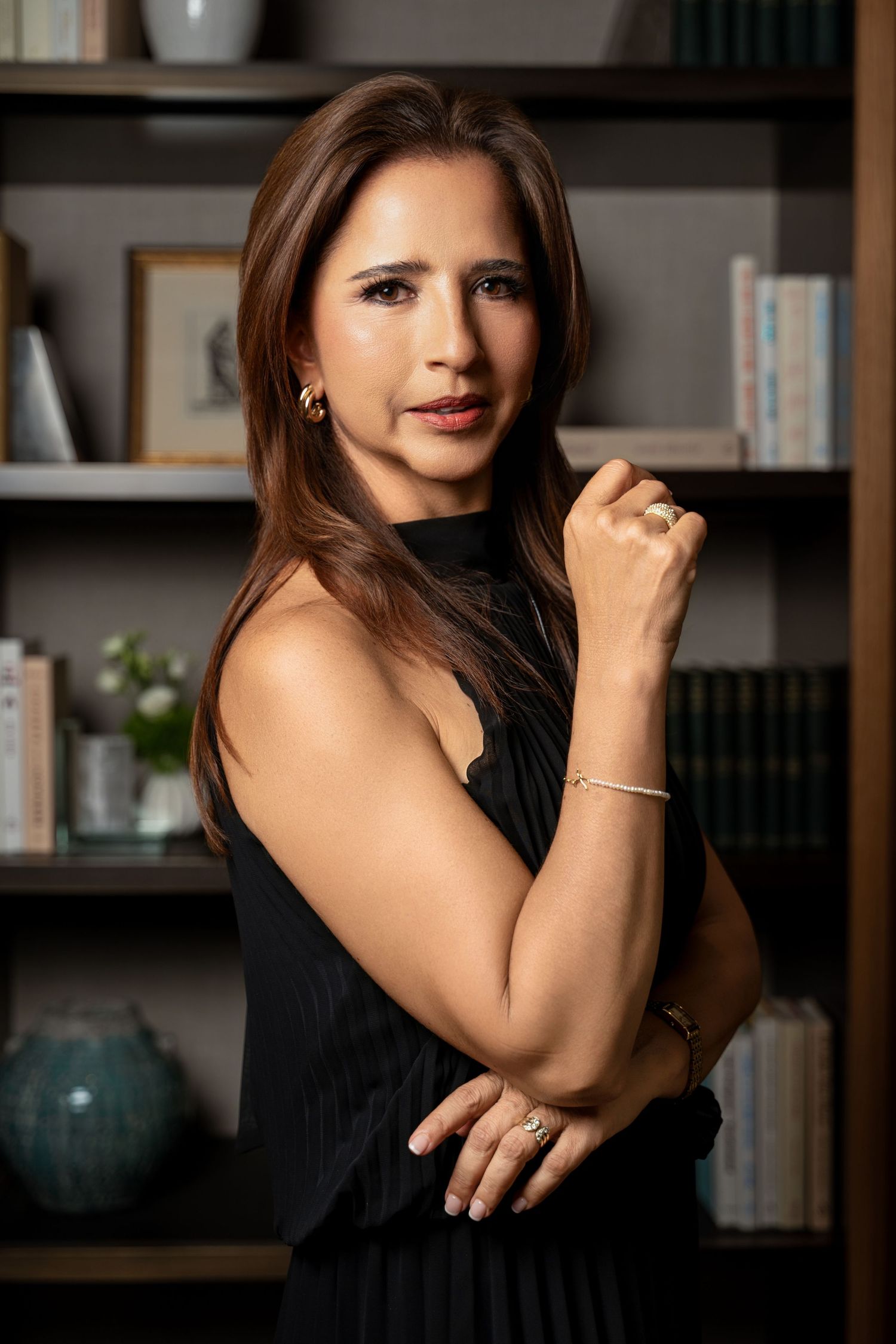 Woman in black dress posing confidently in front of bookshelf, wearing gold jewelry and bracelet.