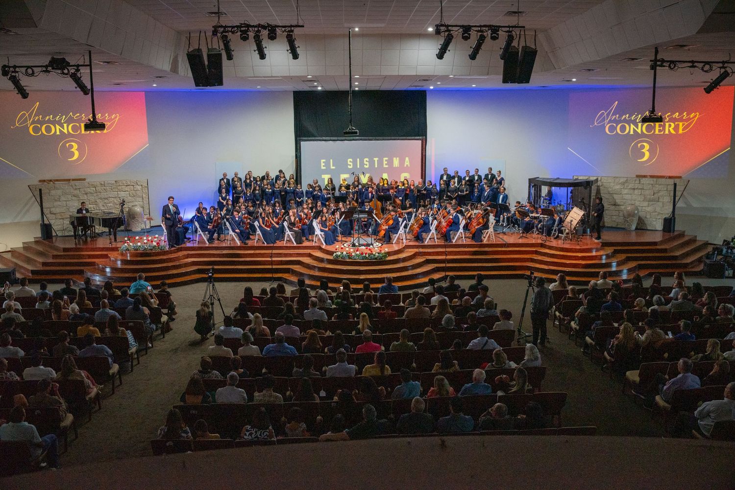 Large orchestra and choir perform on illuminated stage with curved wooden risers in front of seated audience.