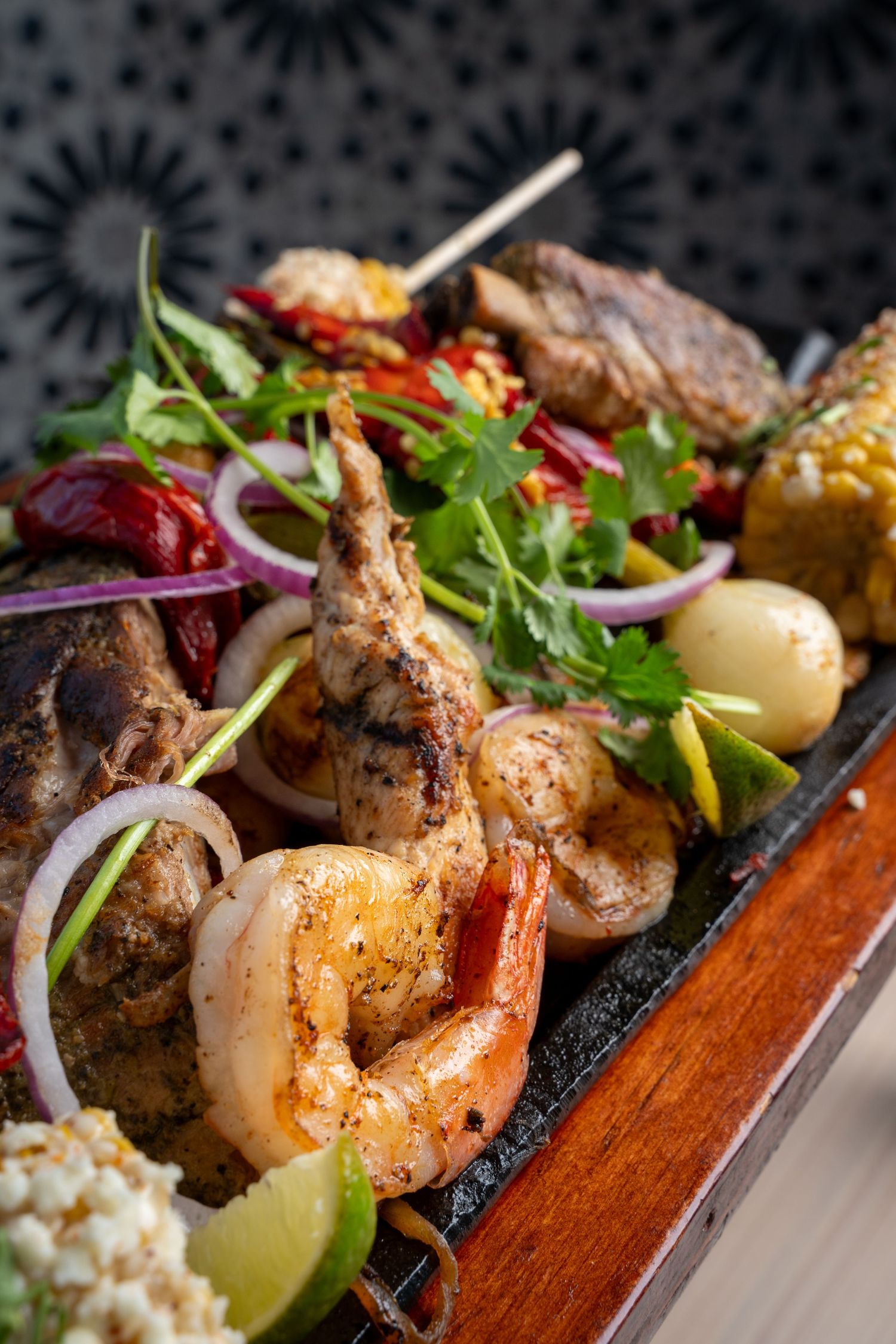 Grilled meat and seafood kebabs with colorful vegetables and corn on a wooden serving board.