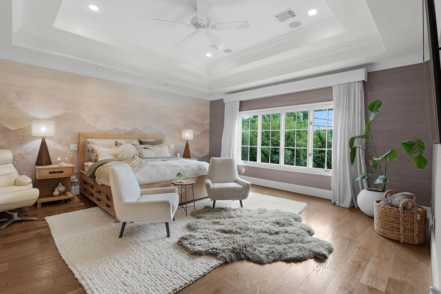 Elegant master bedroom with tray ceiling, hardwood floors, plush area rug, neutral tones, and large window with garden view.