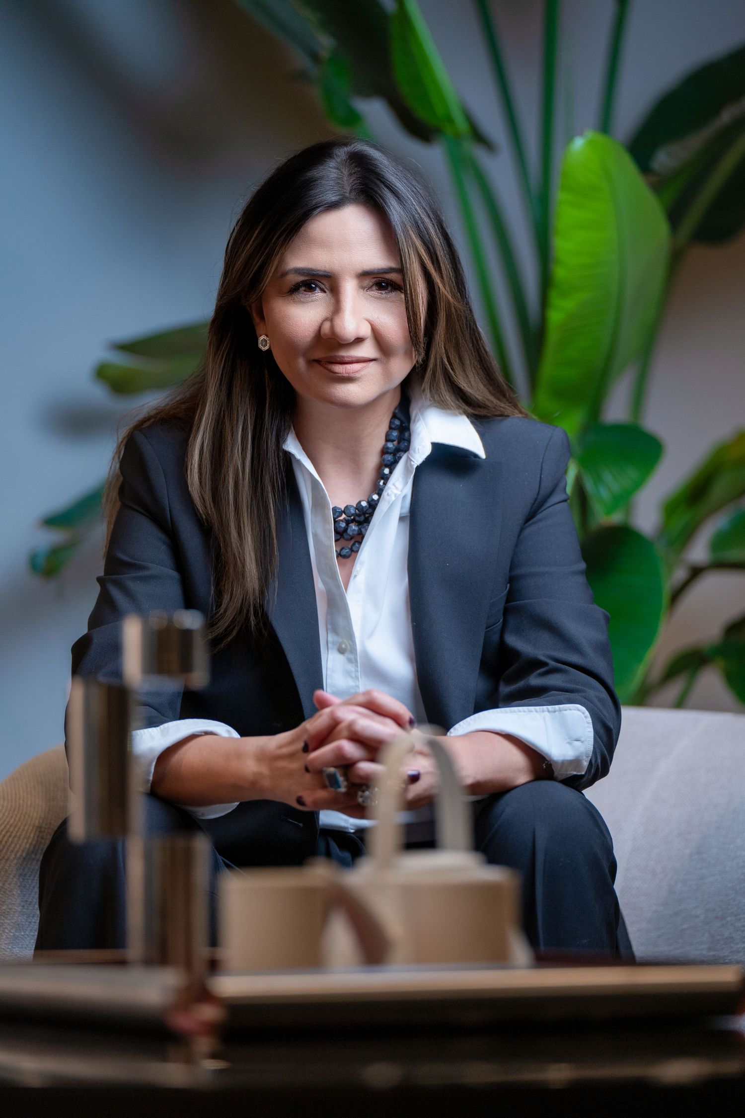 Professional in business attire sits on couch with coffee table in foreground and green plant leaves in background.