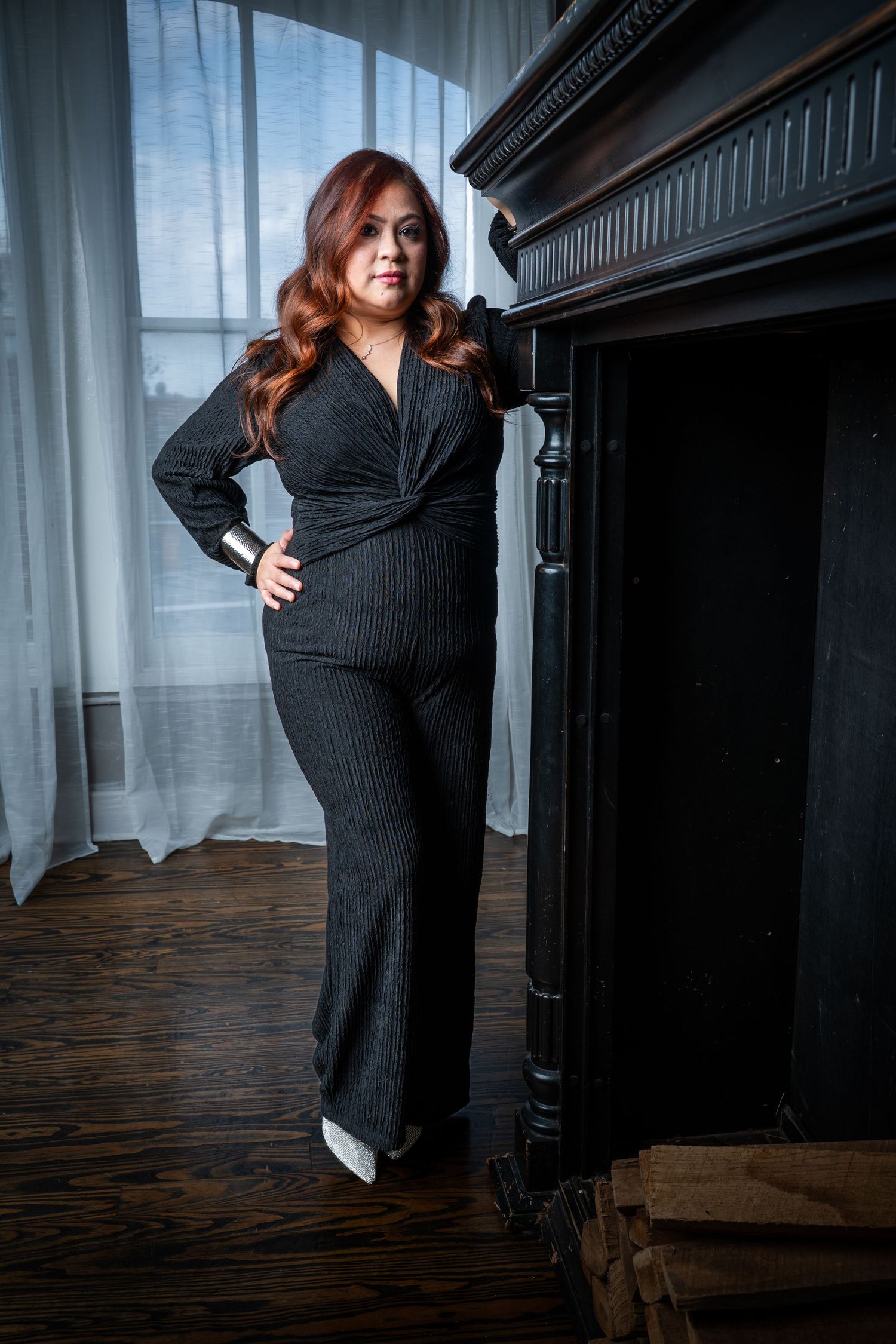 A person in a black dress poses gracefully near a piano in a dimly lit room with wooden floors and curtains.