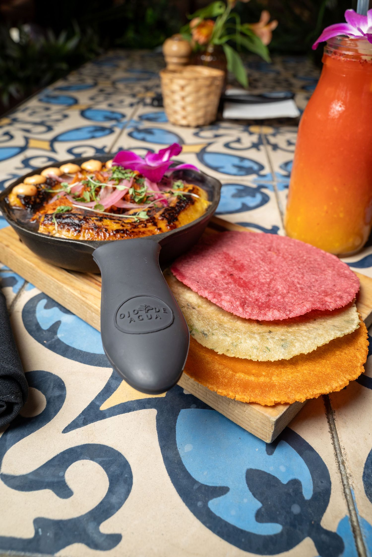 Cast iron skillet with garnished dish and colorful tortillas on decorative tile table.