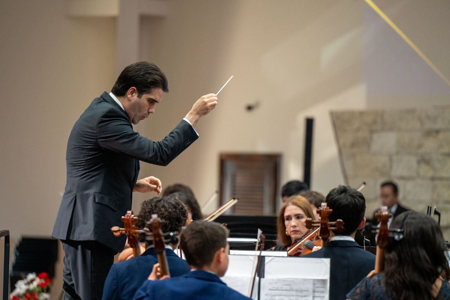 Orchestra conductor in dark suit leads musicians with baton during classical performance.