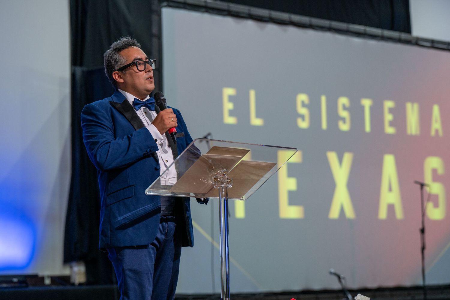 Speaker presenting at a conference podium with yellow text displaying 'EL SISTEMA TEXAS' on the screen behind.