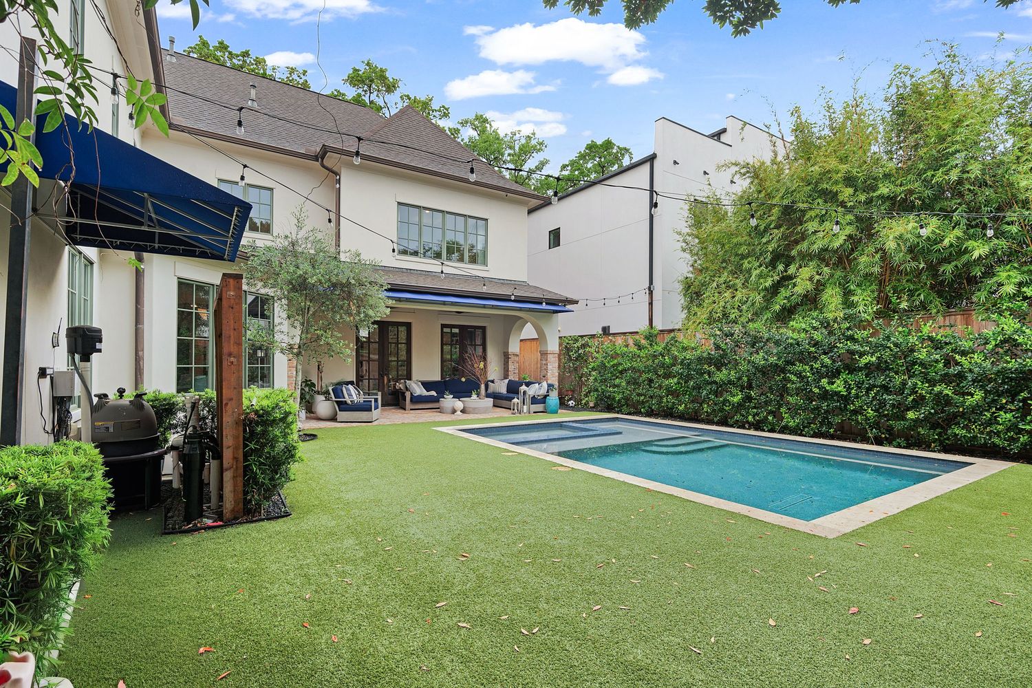 Backyard oasis featuring a rectangular pool, lush green lawn, and a two-story home with a covered patio and blue awning.