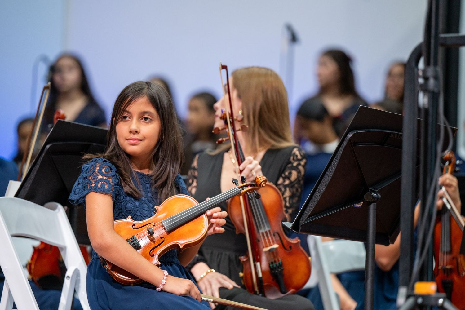 Orchestra musicians with string instruments sit at music stands during a performance in blue evening light.