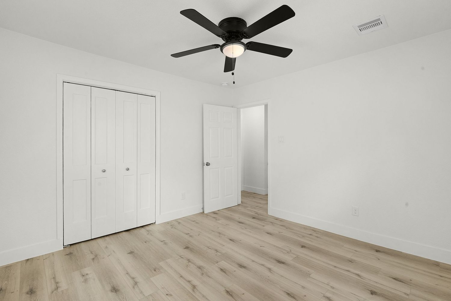 Empty bedroom with light wood flooring, white walls, and a black ceiling fan.