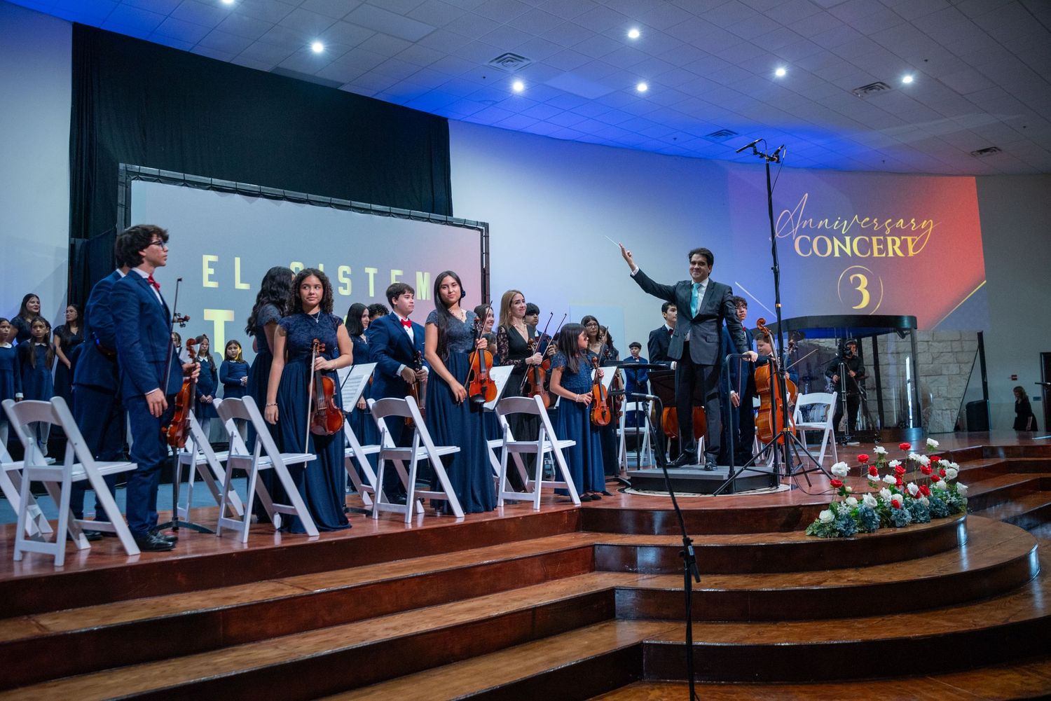 Orchestra ensemble in formal attire performs on stage with blue lighting and tiered seating arrangements.