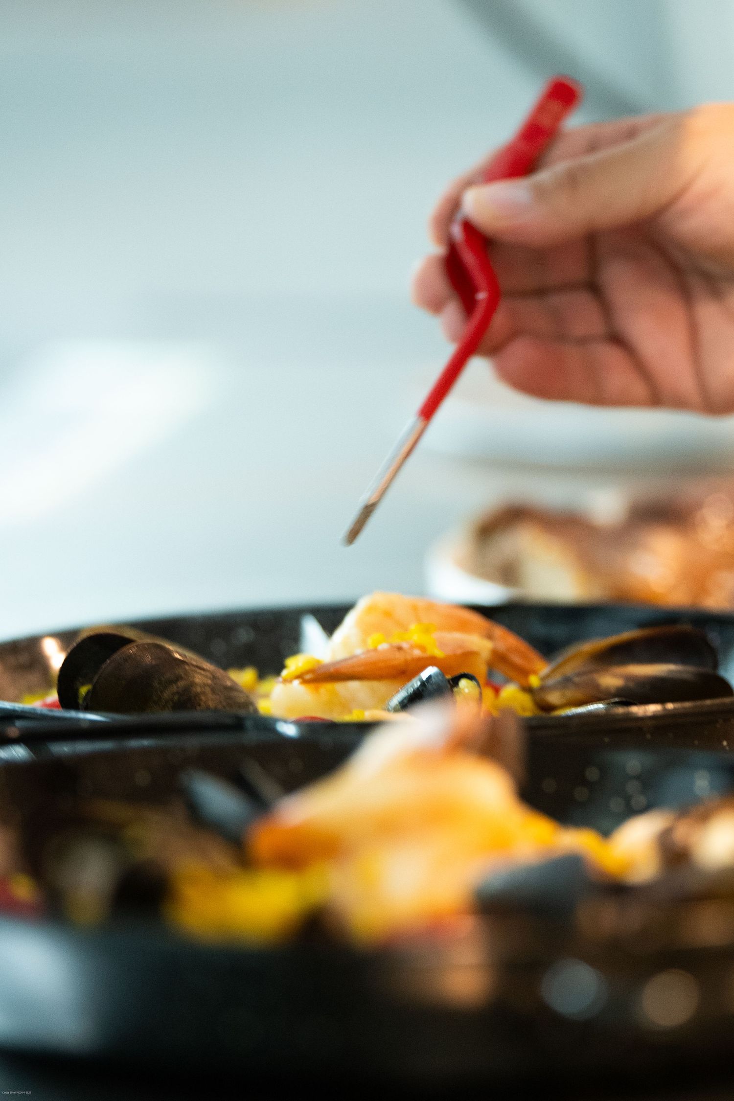 A hand with chopsticks reaches toward a blurred Asian dish on a dark plate with vibrant yellow and orange colors.