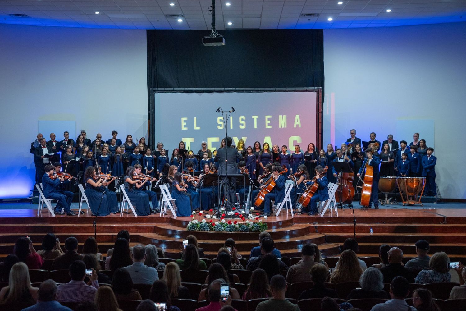 A full symphony orchestra performs on stage with string and wind sections beneath a projection screen displaying 'El Sistema'.