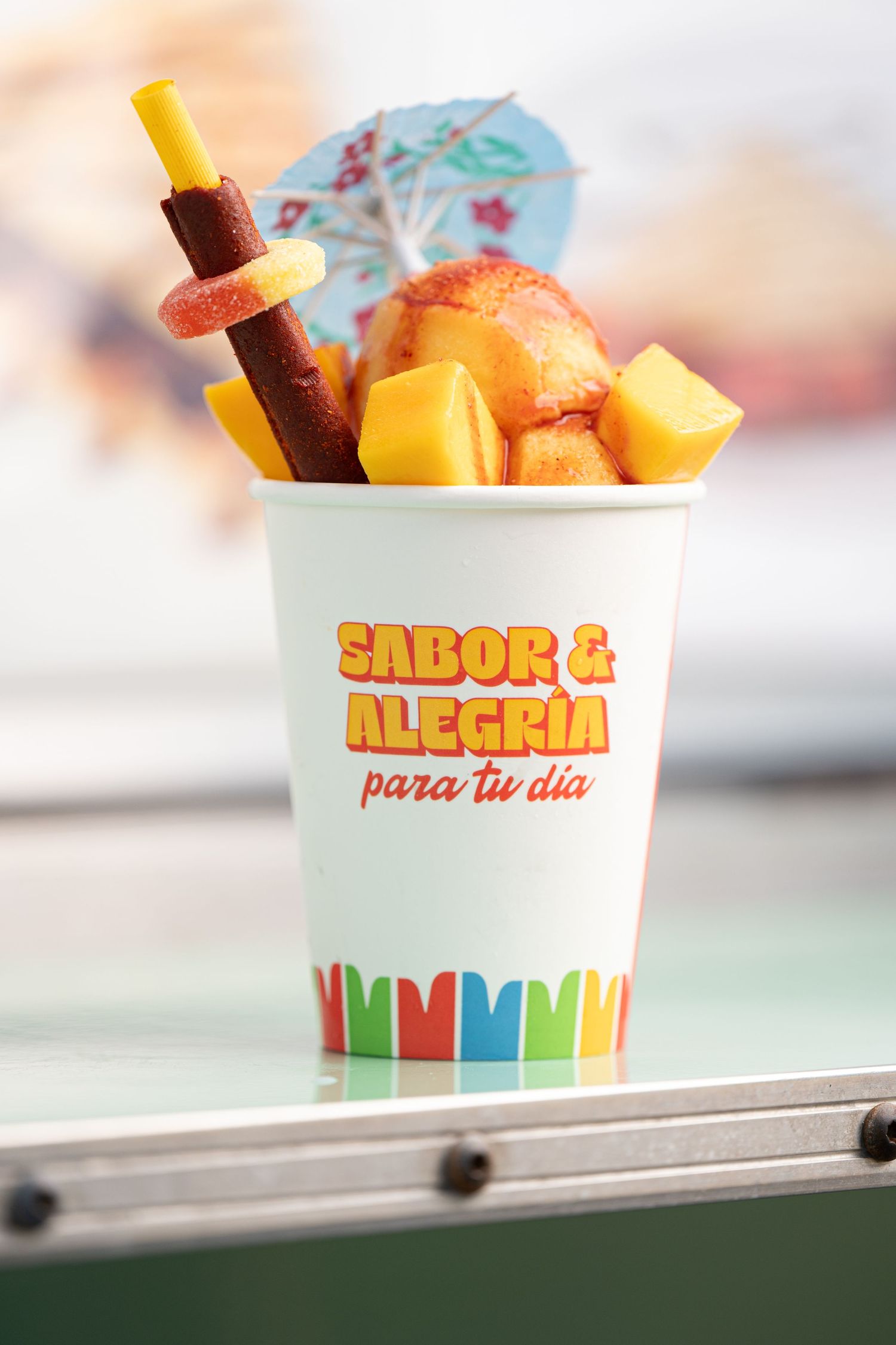 A colorful drink cup labeled 'Sabor & Alegria' filled with fresh fruit chunks and a garnish under a blue umbrella.