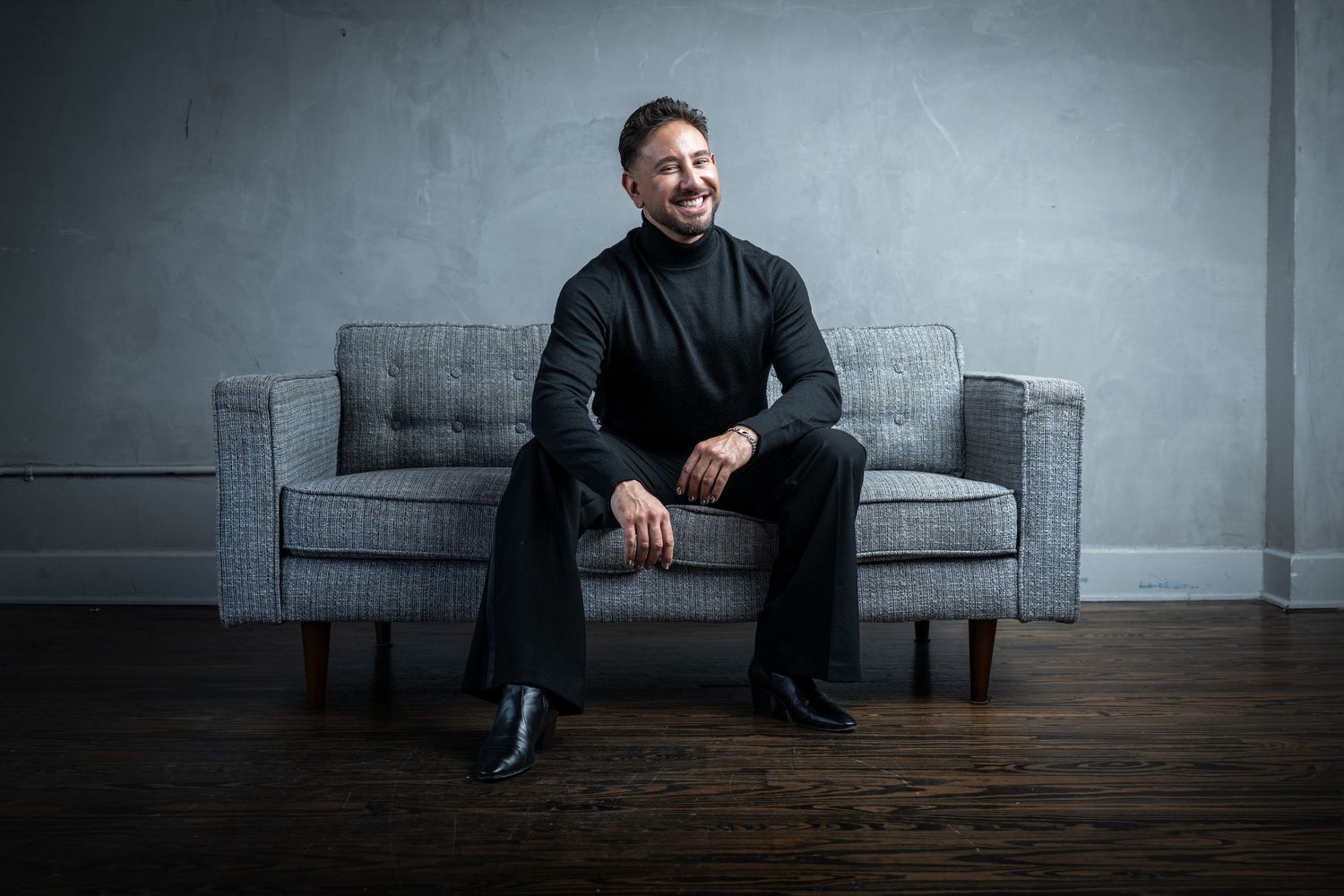 A figure sits on a modern gray couch in a minimalist room with hardwood floors and gray walls.