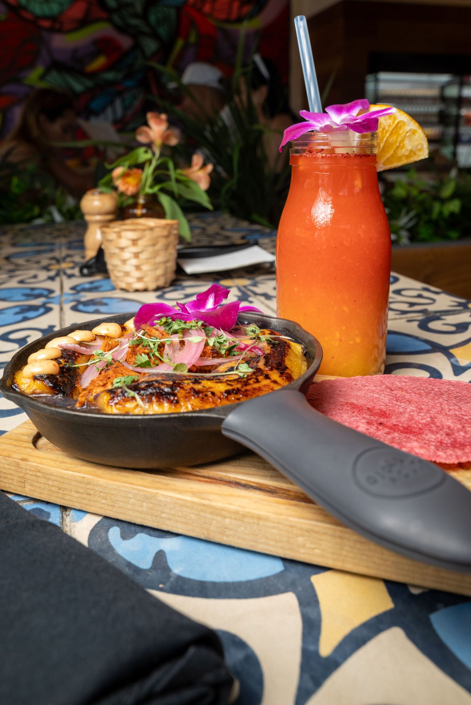 Orange tropical smoothie and garnished skillet dish on blue patterned table setting.