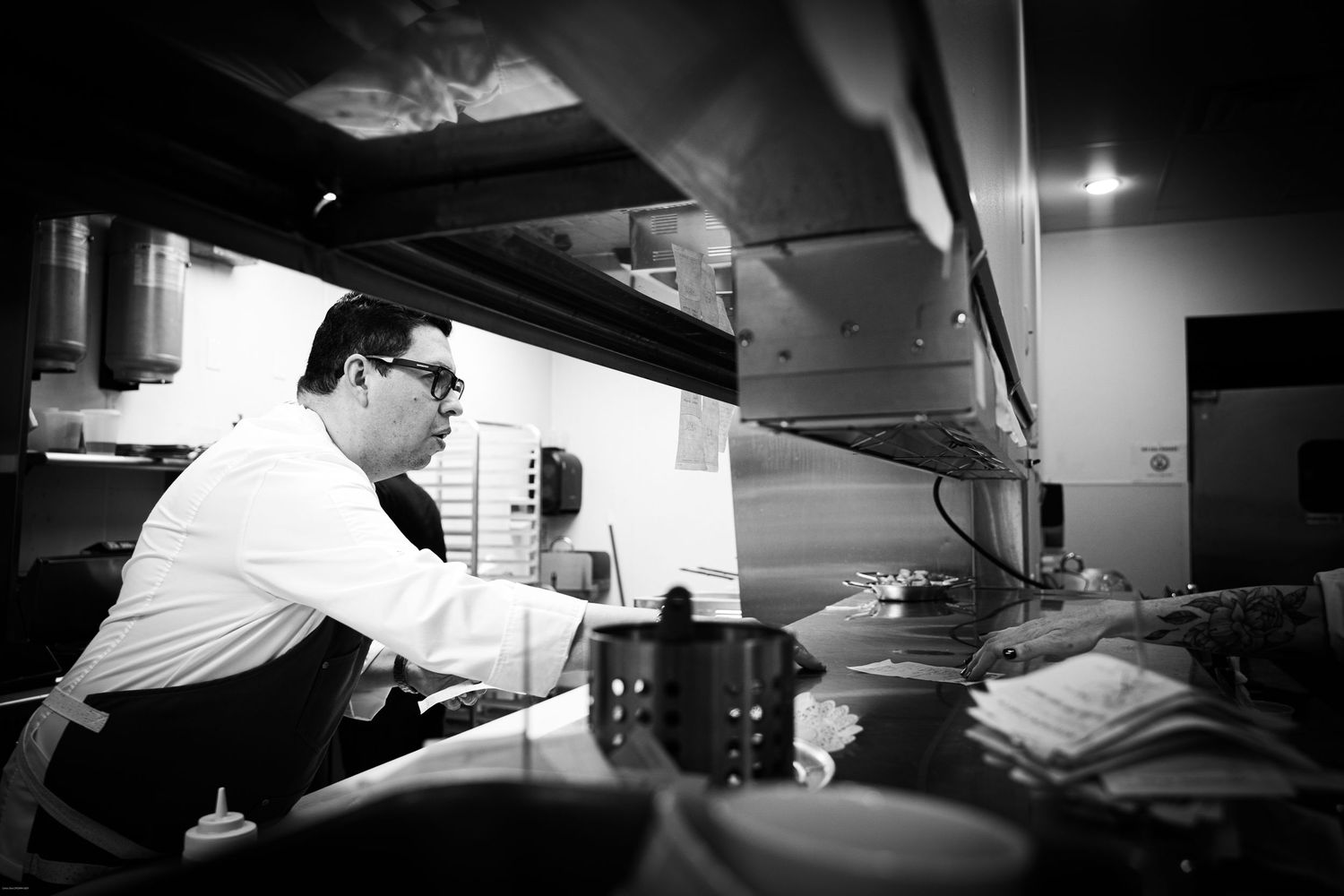 A chef in white works at a busy restaurant kitchen station in black and white photography.