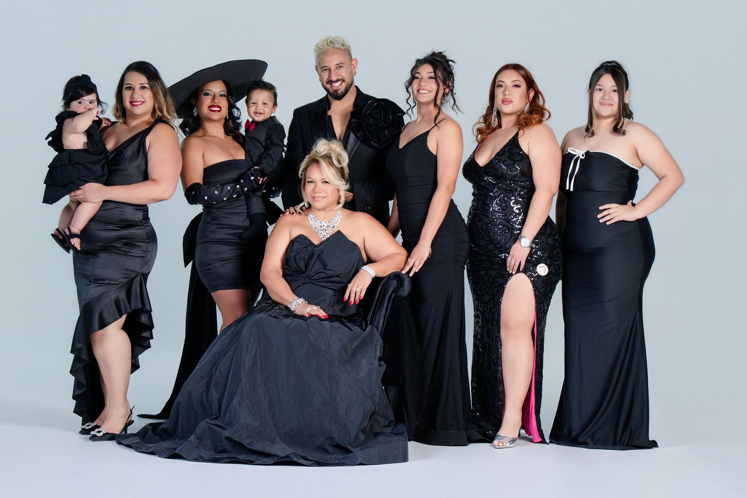 A group of women pose together wearing elegant black formal dresses and gowns against a light gray background.