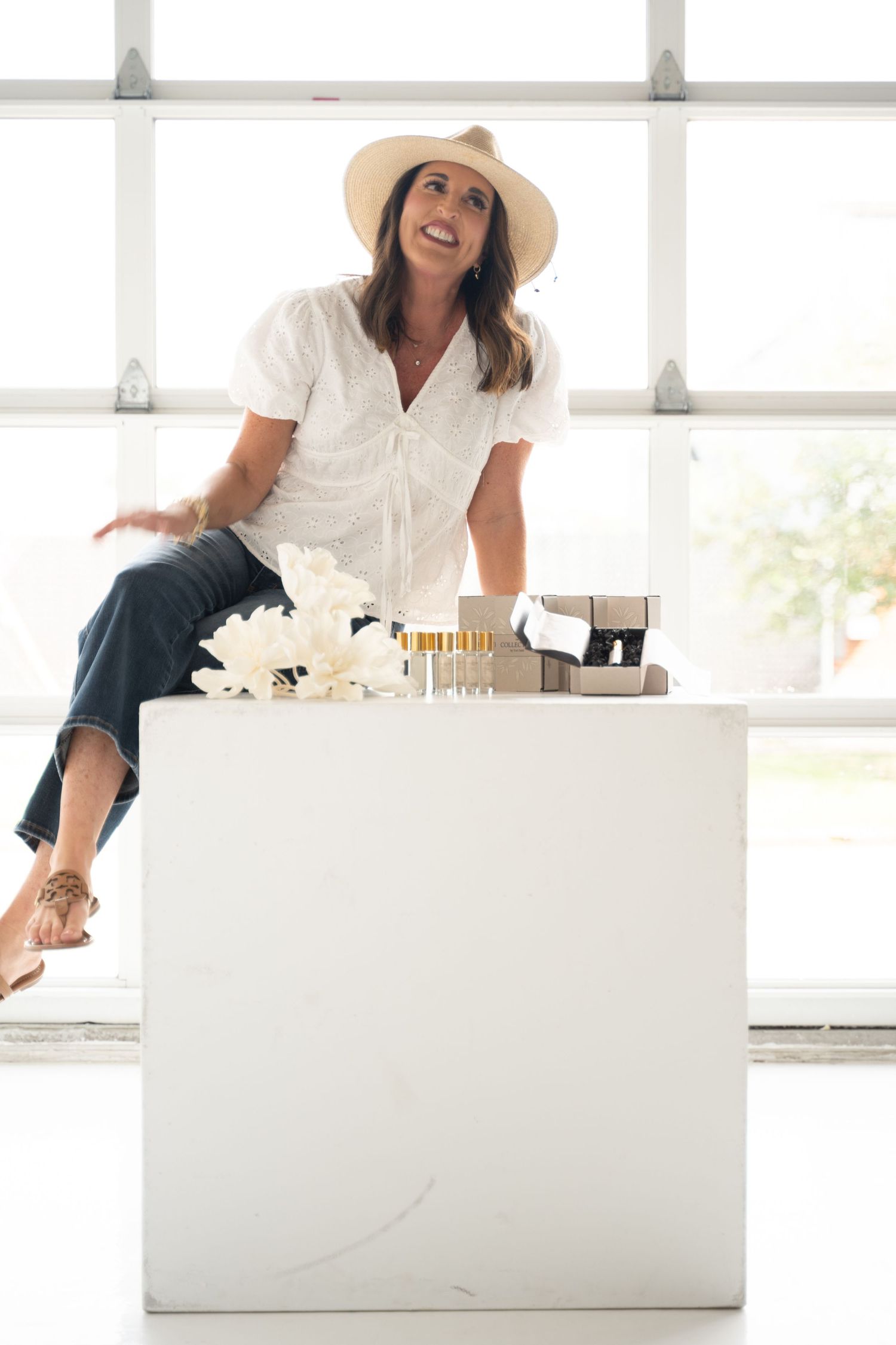 Woman in white blouse and sun hat sitting on white counter with perfume bottles and white flowers.