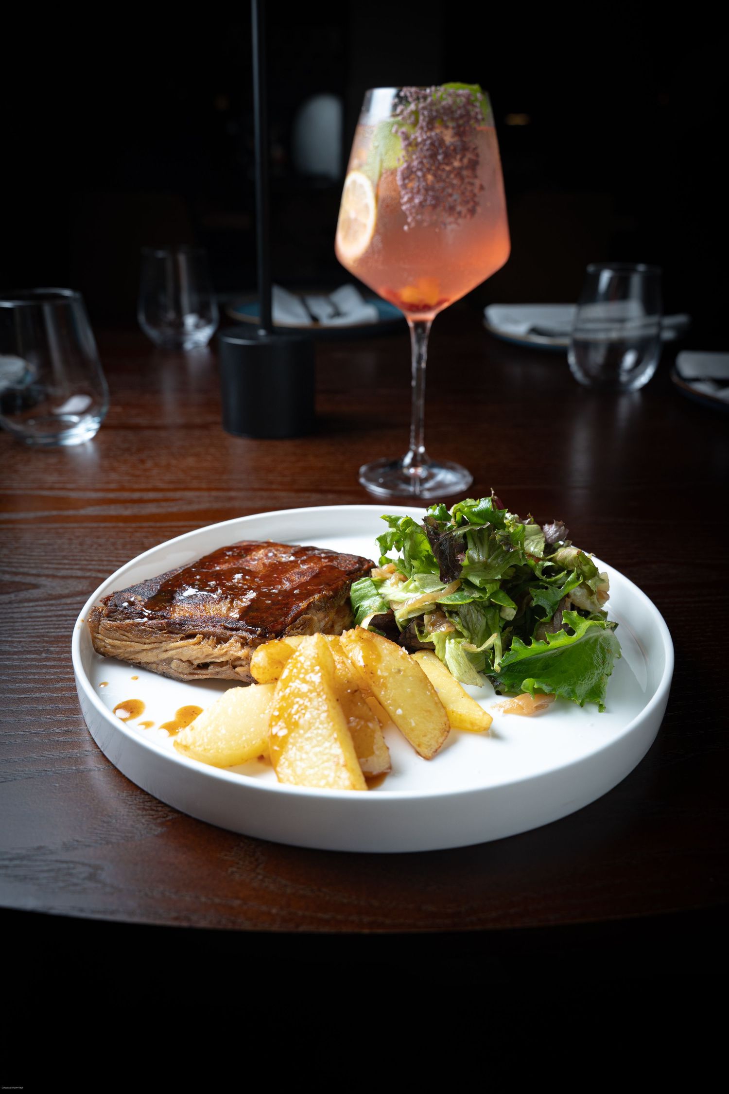 A series of plated dishes and cocktails on a dark wooden table in moody restaurant lighting.