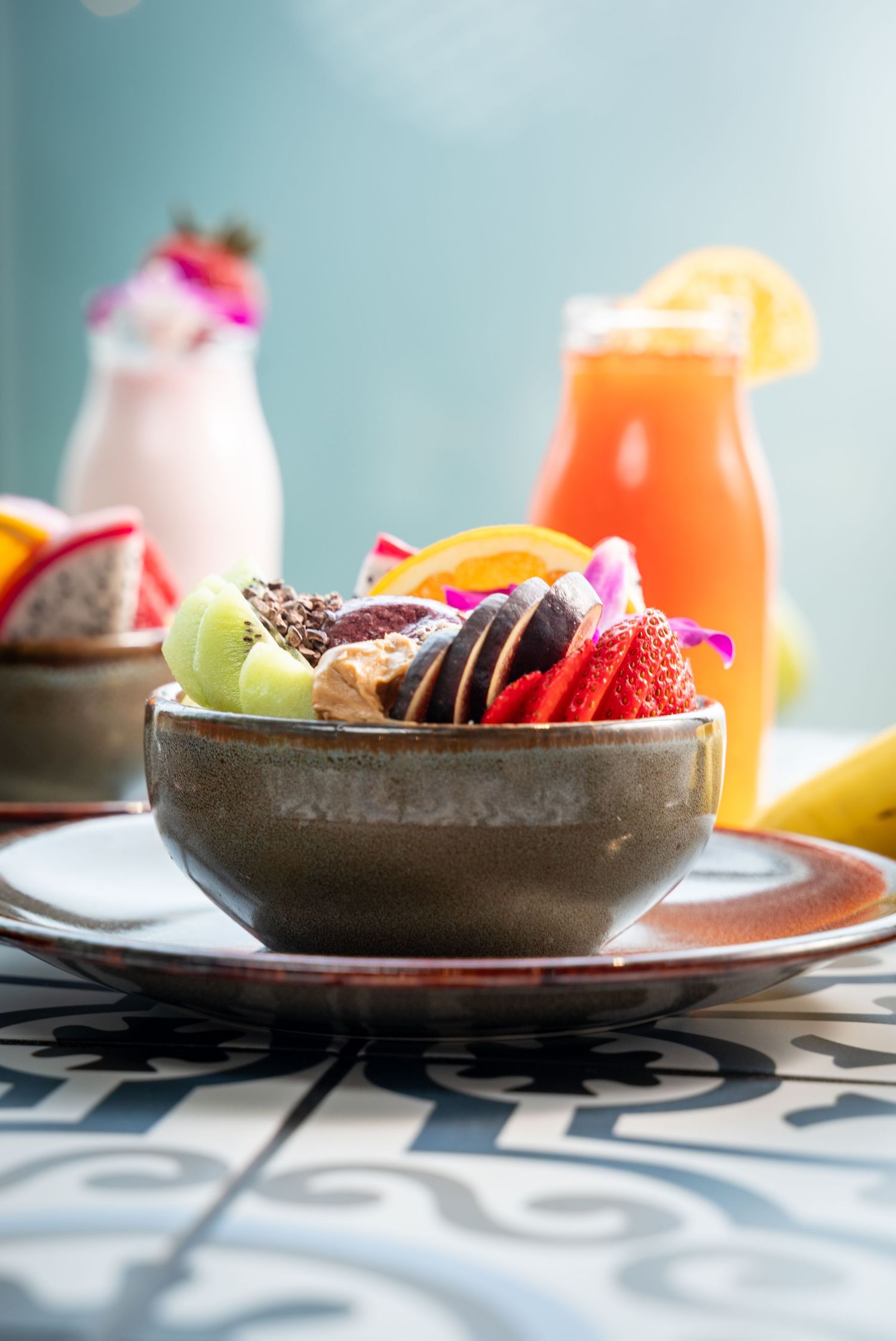 Bowl of fresh cut fruit with smoothies and pink flowers on patterned surface.