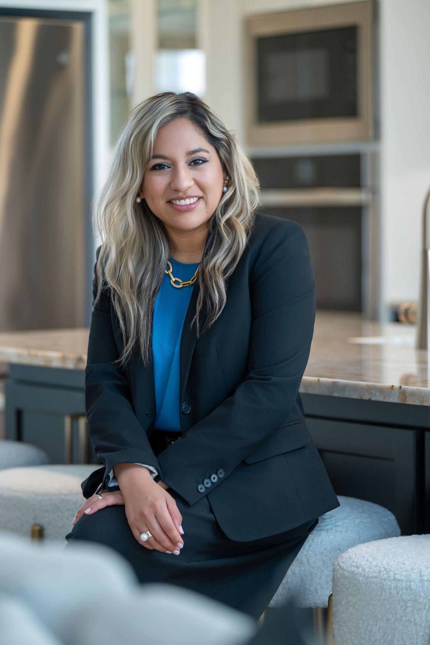 Professional wearing a black blazer and blue top sits casually on modern office furniture in a bright interior setting.