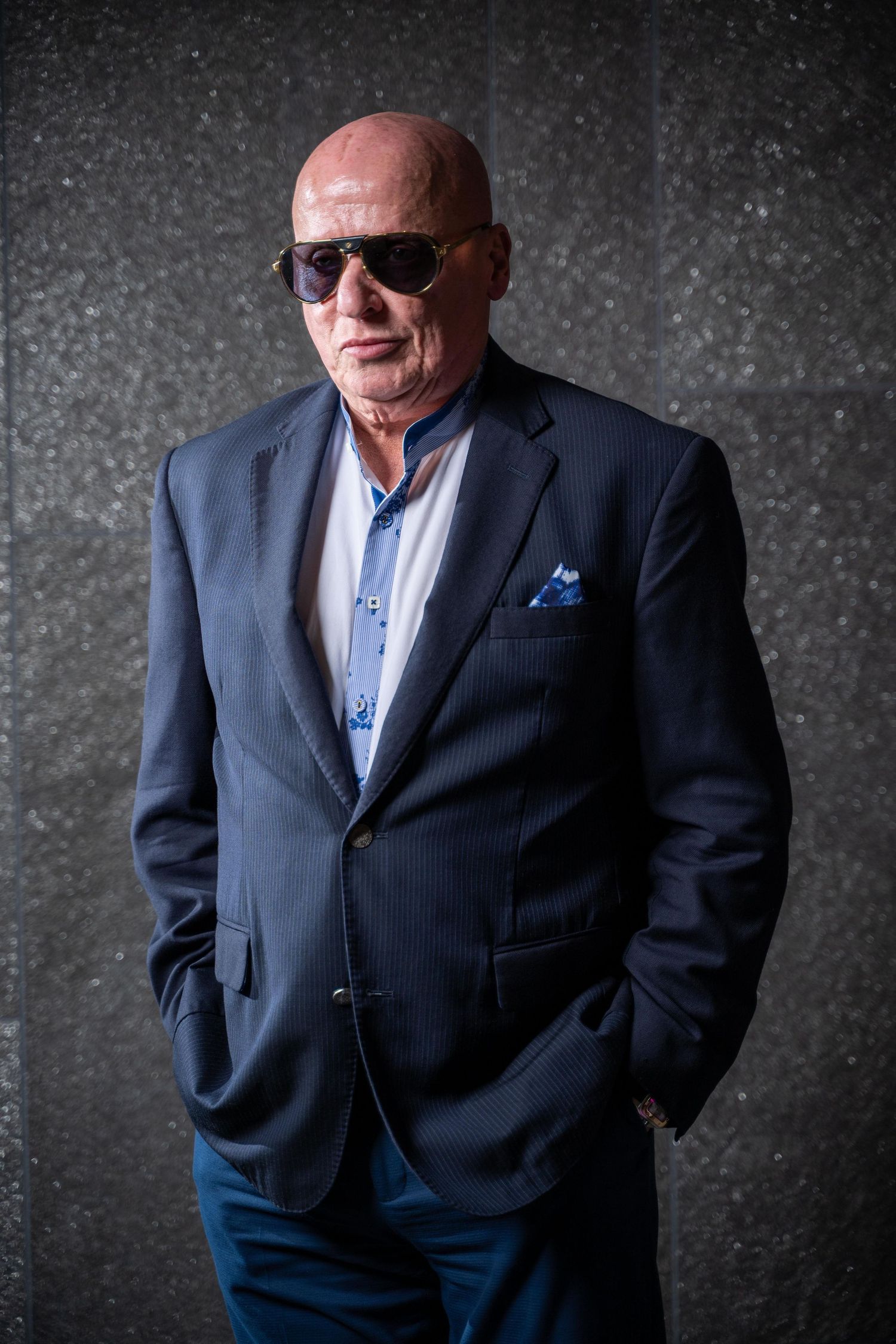 A distinguished individual in a navy blazer, sunglasses and blue shirt poses against a textured dark background.