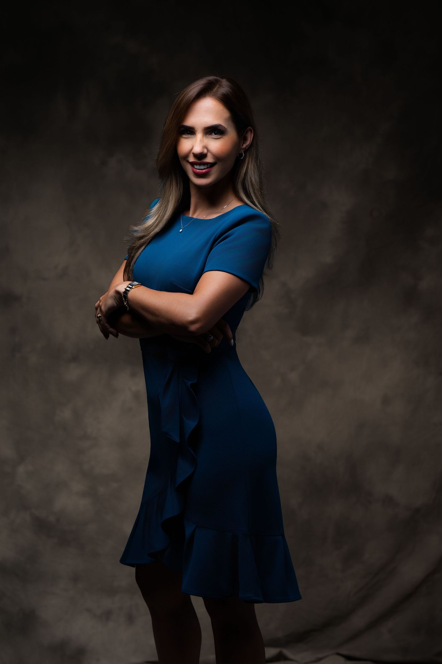 A woman in a fitted navy blue dress poses confidently with arms crossed against a dark dramatic background.