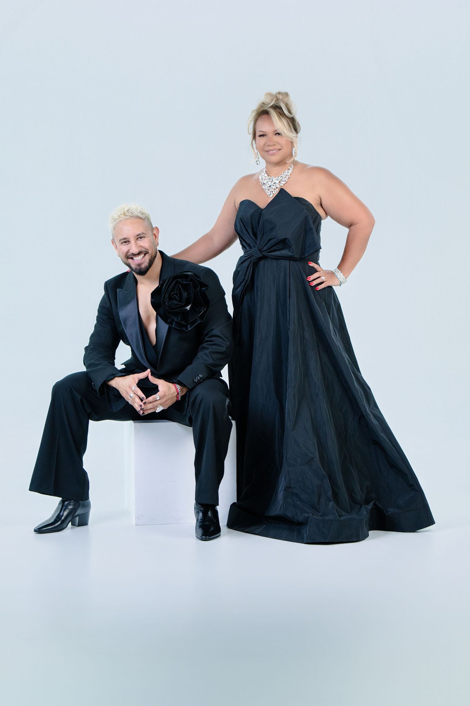 A couple in elegant formal black attire pose together against a white background for a glamorous photo shoot.