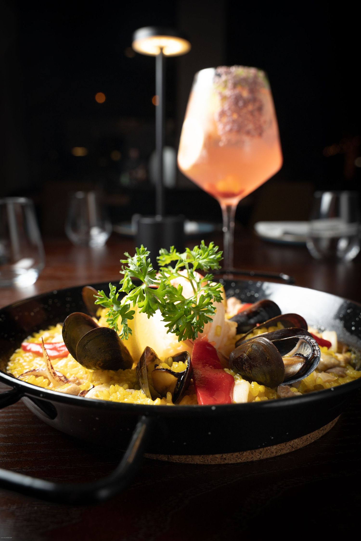 A seafood paella dish in a cast iron skillet with mussels and fresh herbs, accompanied by a pink cocktail in a wine glass.