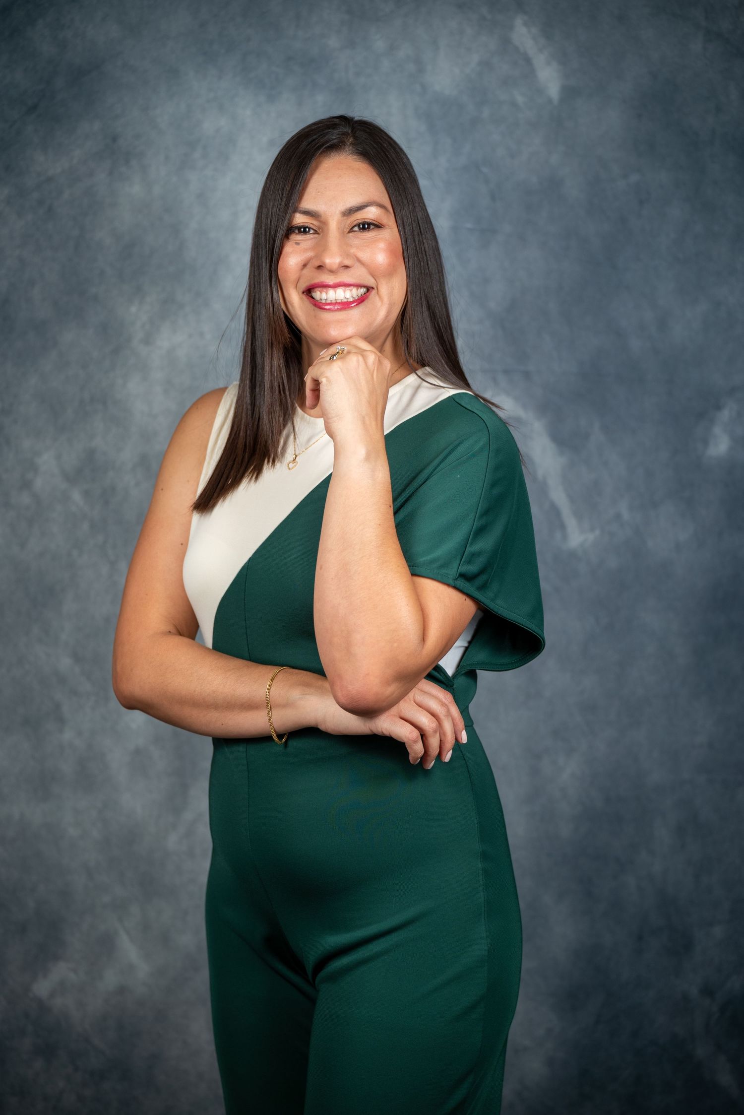 Smiling woman in green and white jumpsuit poses confidently against gray backdrop in professional portrait.