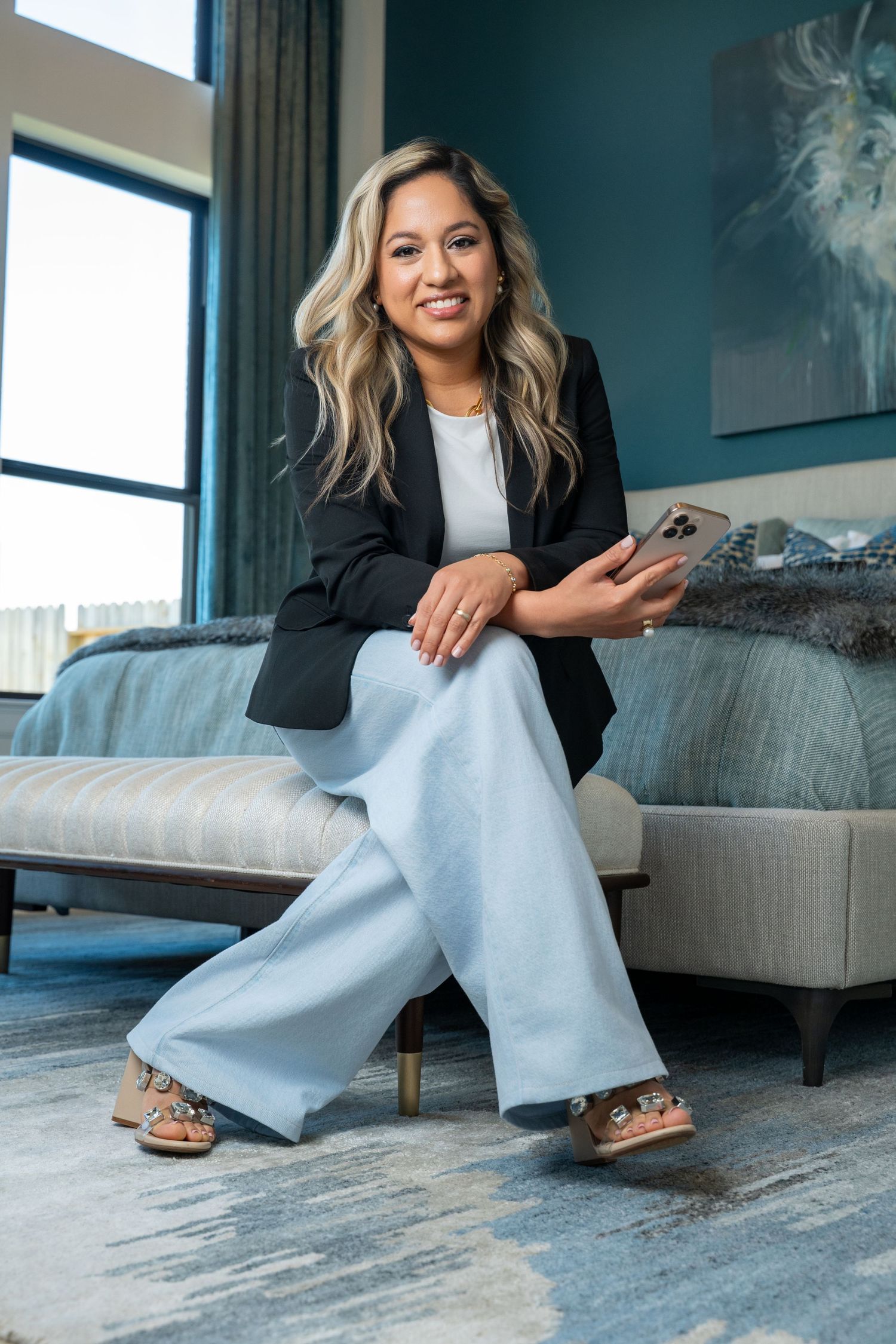 Woman in black blazer and light blue wide-leg pants sits on bench holding phone in elegant teal bedroom.