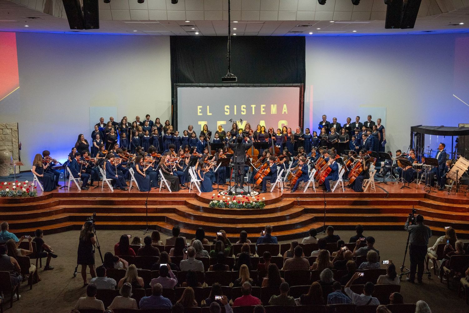 Large orchestra and choir perform on illuminated stage with curved wooden risers in front of seated audience.