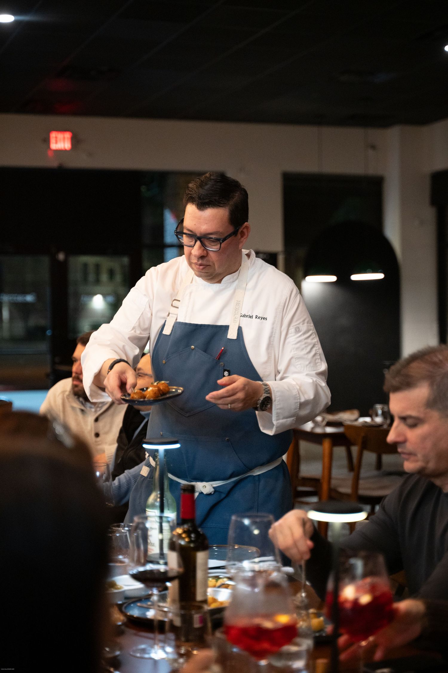 A chef in white uniform and blue apron serves food to diners at a dimly lit restaurant table.