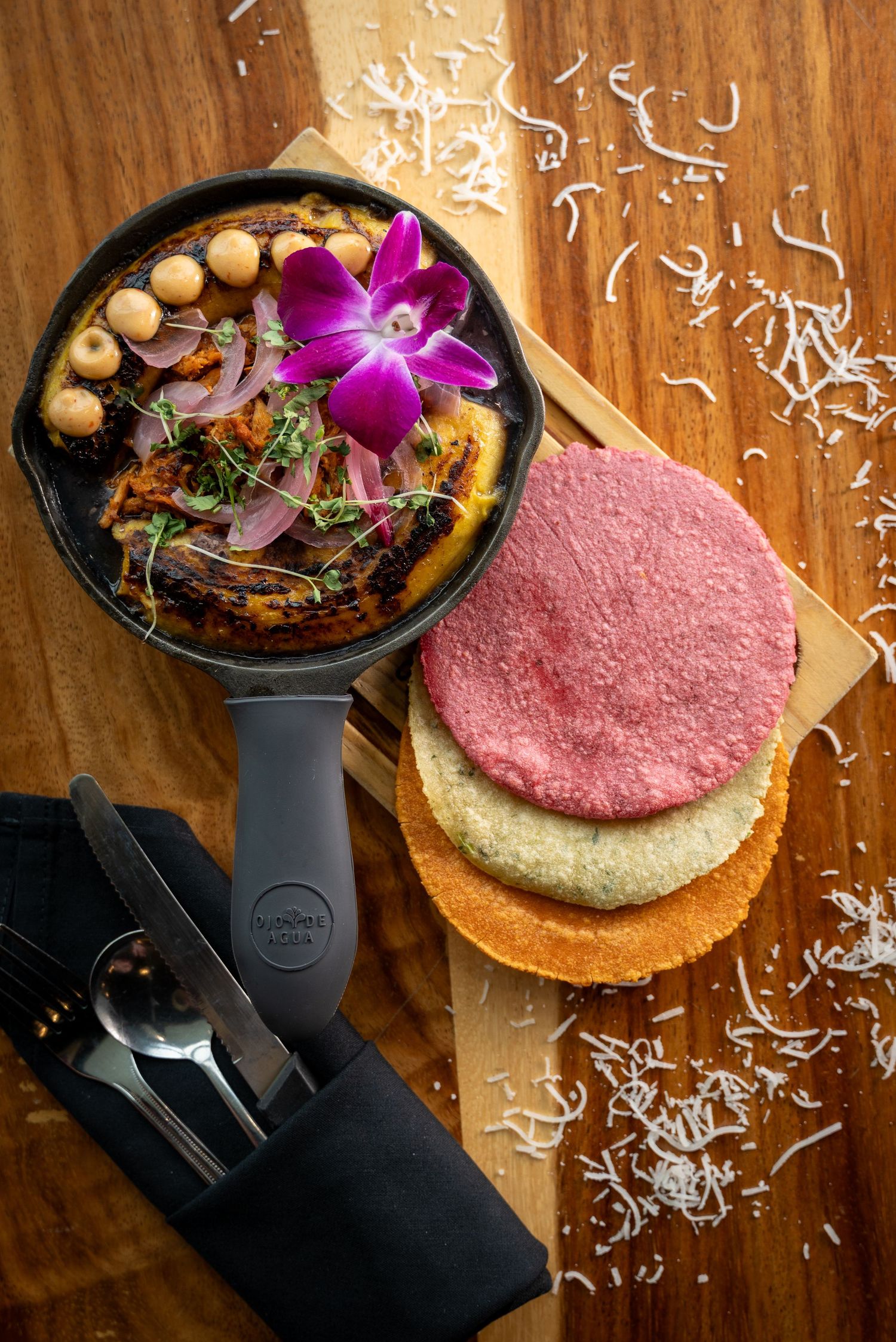 Black serving pan with colorful salad decorated with purple orchid and three colorful tortillas on wooden surface.