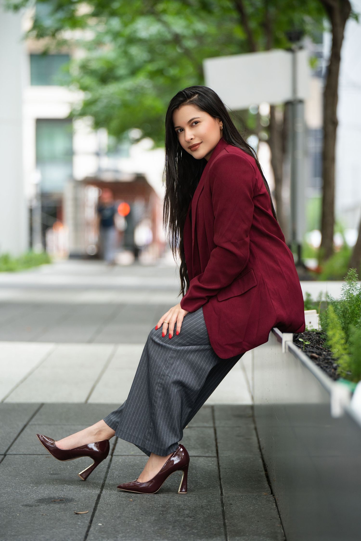 Professional fashion portrait of a person in a burgundy blazer and gray skirt posing on an urban sidewalk.
