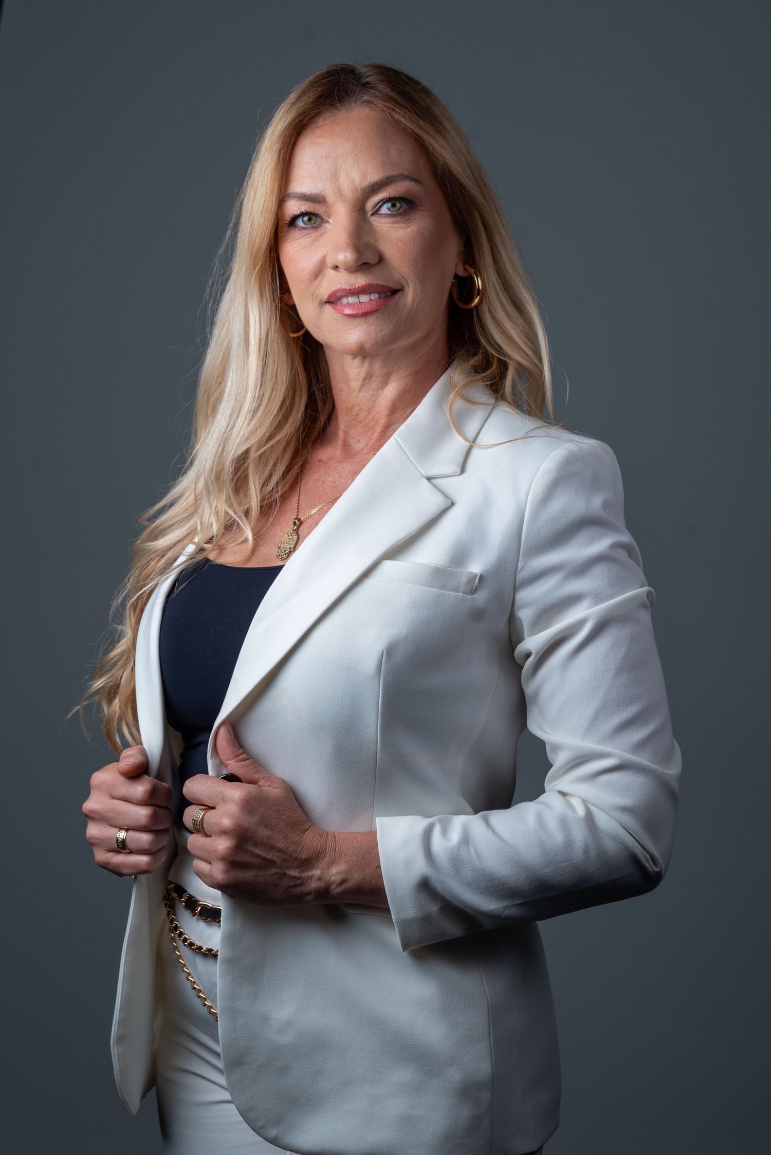 Professional blonde woman in white blazer posing confidently against gray background for business portrait.