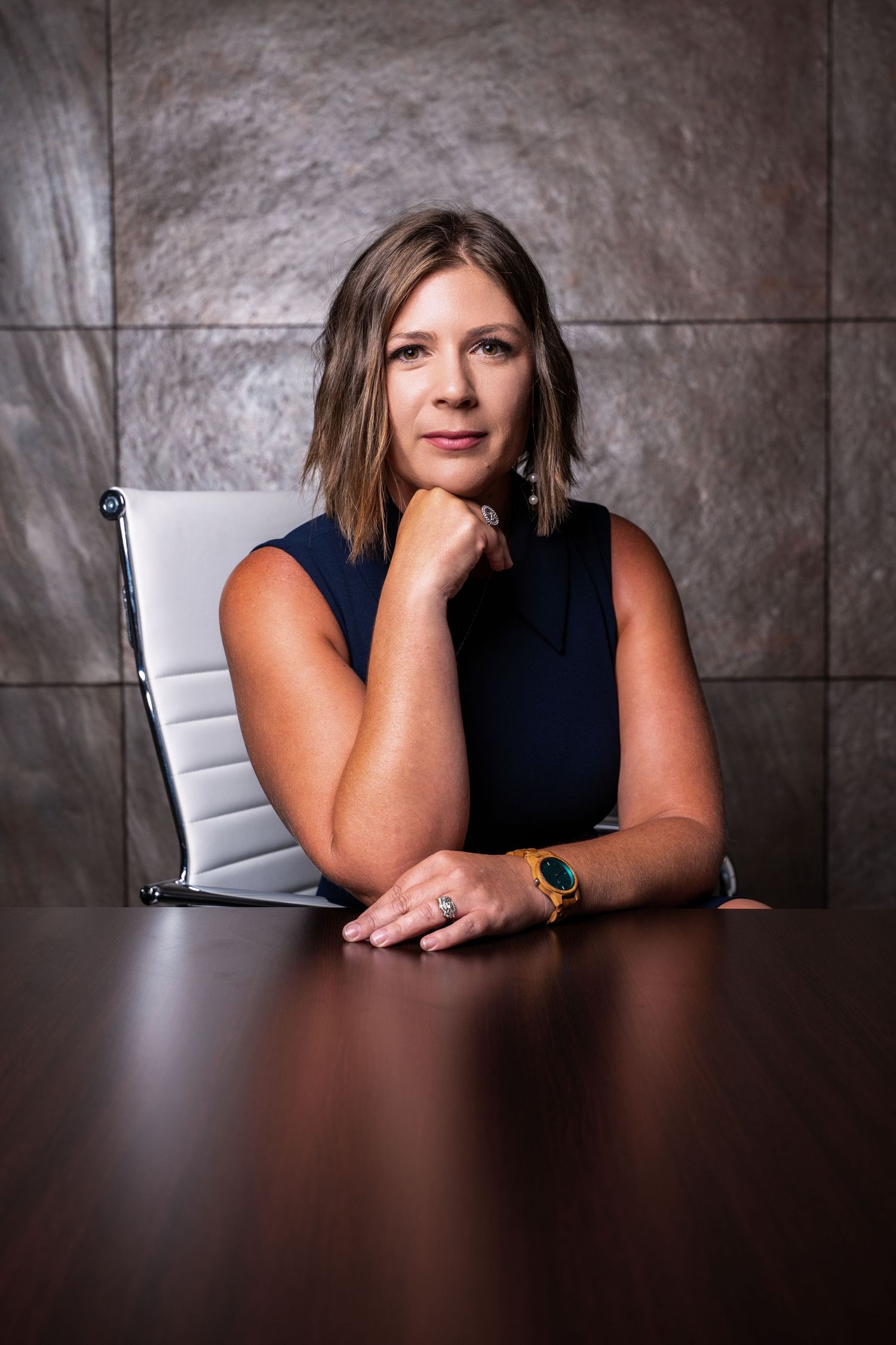 Professional portrait of someone in a navy sleeveless top seated at a dark wood table against a textured gray backdrop.