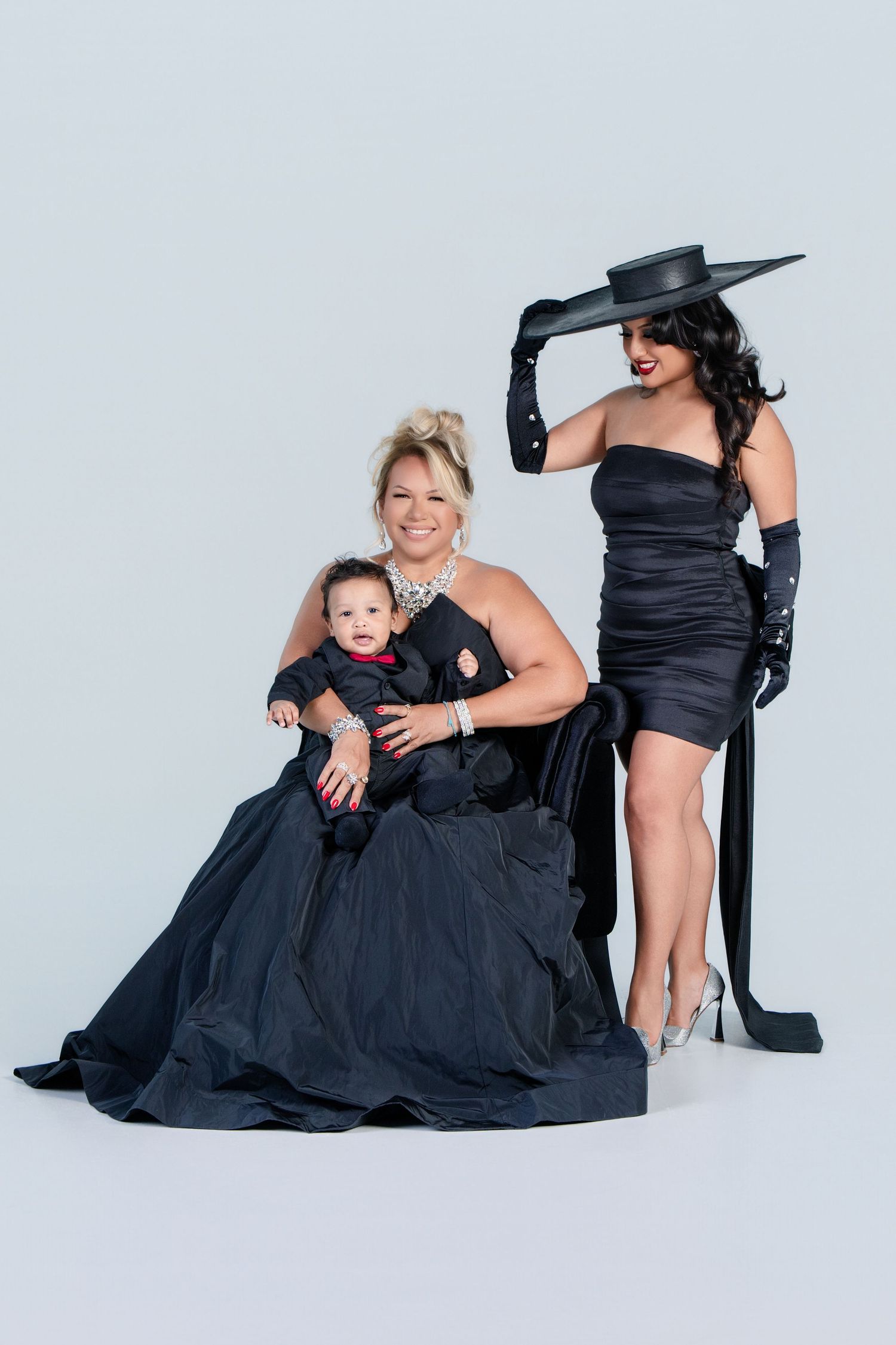 Three generations pose in elegant black formal wear including dresses, gloves and a wide-brimmed hat against a light backdrop.