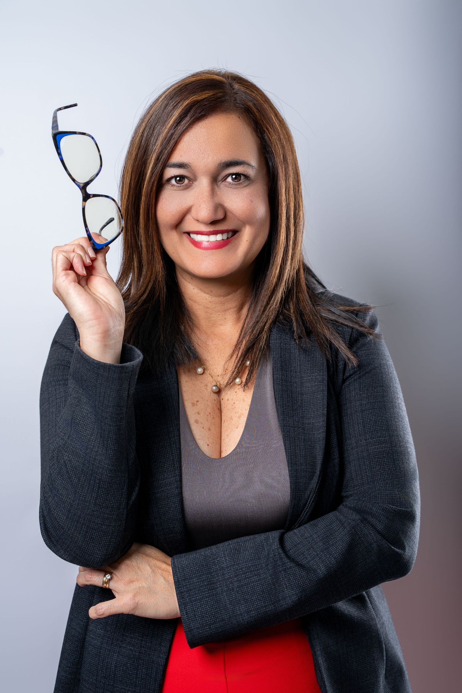 Professional business portrait showing person in dark blazer and red skirt holding eyeglasses with a warm smile.