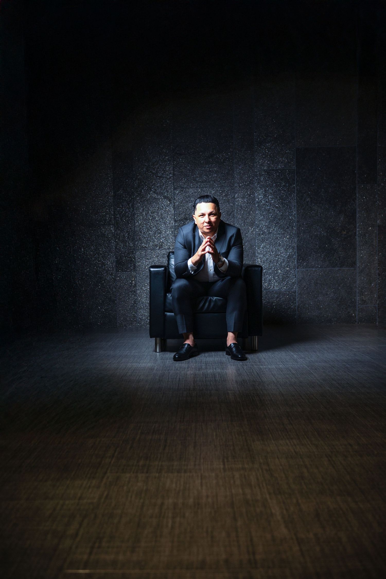 Figure seated in a dark leather armchair against textured wall in moody atmospheric lighting.