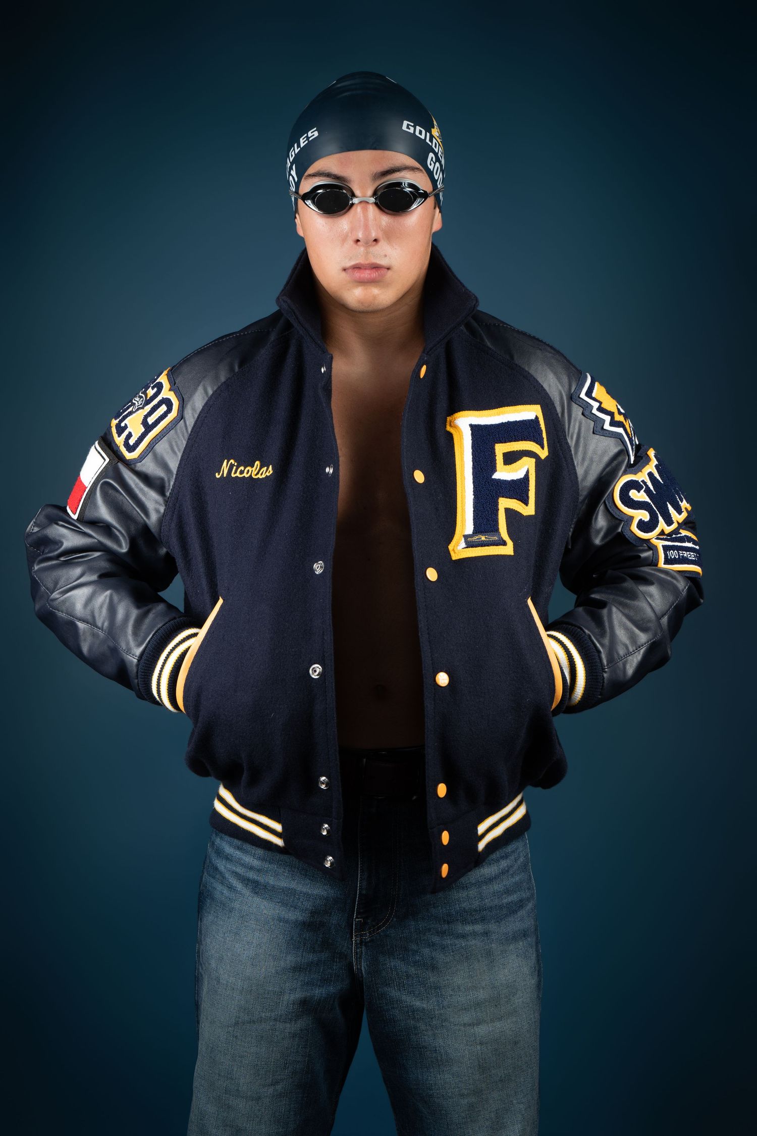 Young man wearing a varsity letterman jacket with swim goggles and cap poses confidently against dark background.