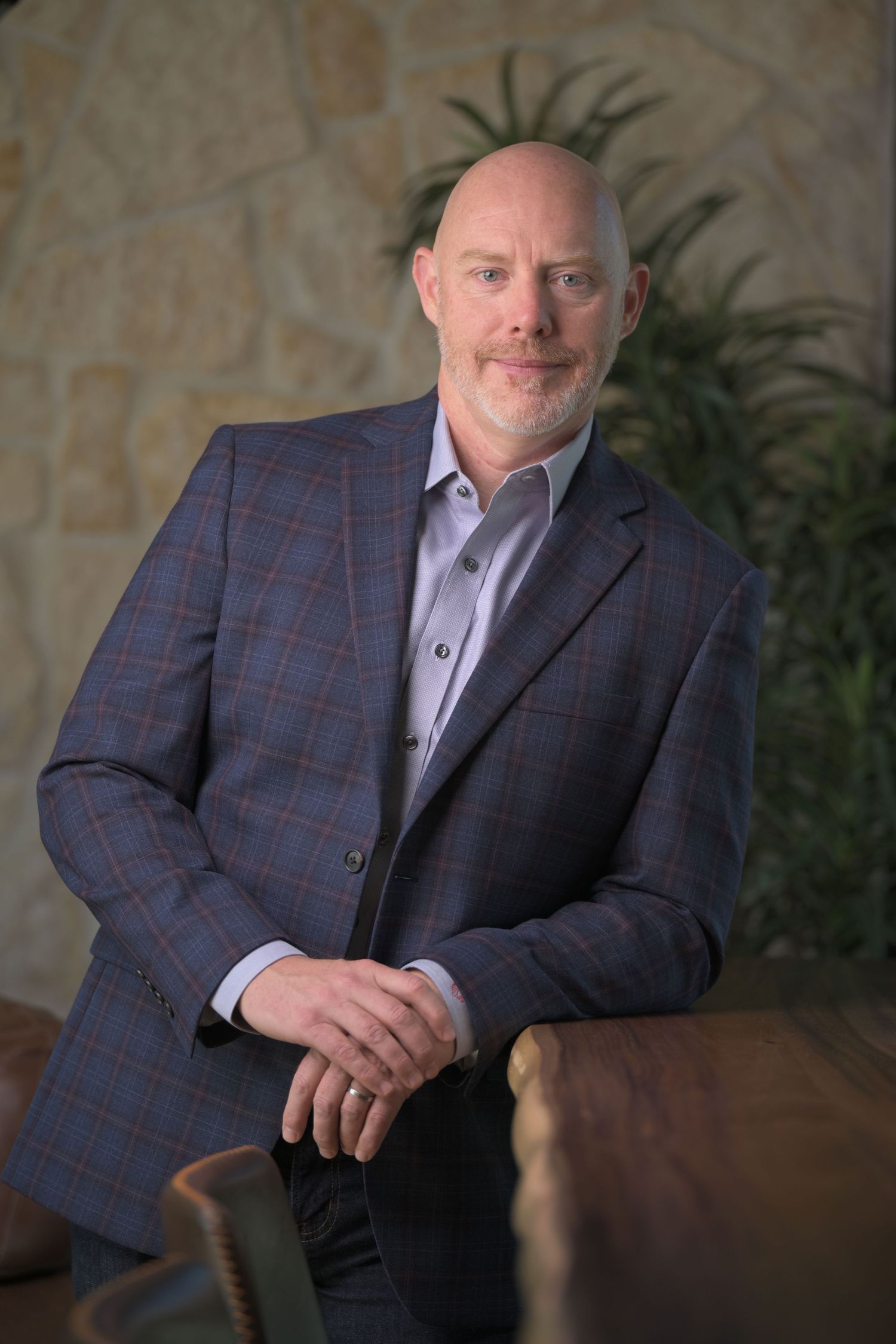 Bald man with beard wearing blue plaid blazer poses confidently in modern office with stone wall background.