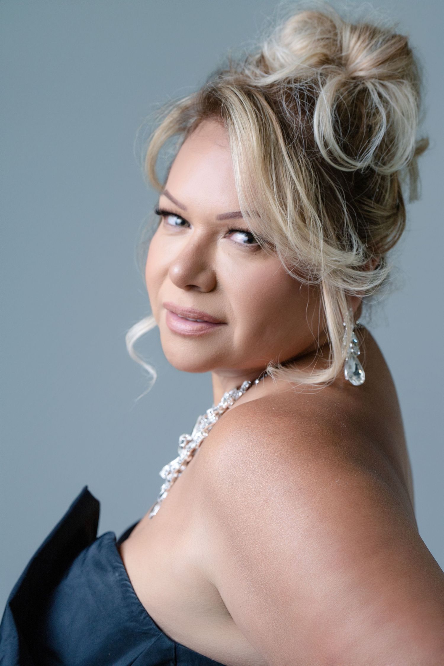Elegant portrait showing blonde updo hairstyle with loose curls, sparkling crystal necklace and navy dress against gray background.