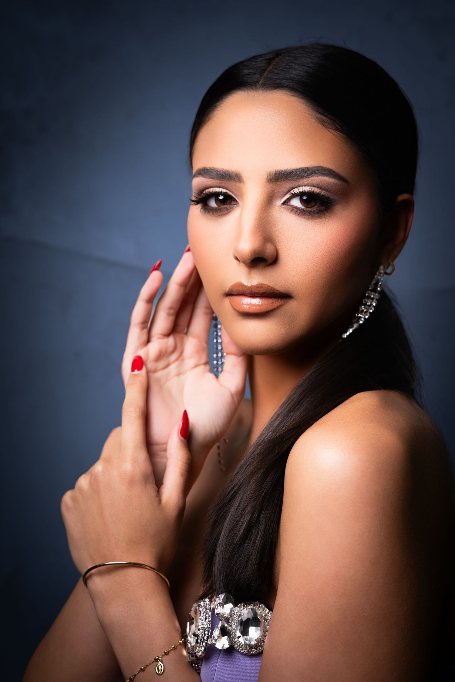 Studio portrait with dramatic lighting showcases glamorous makeup and jewelry against dark background.